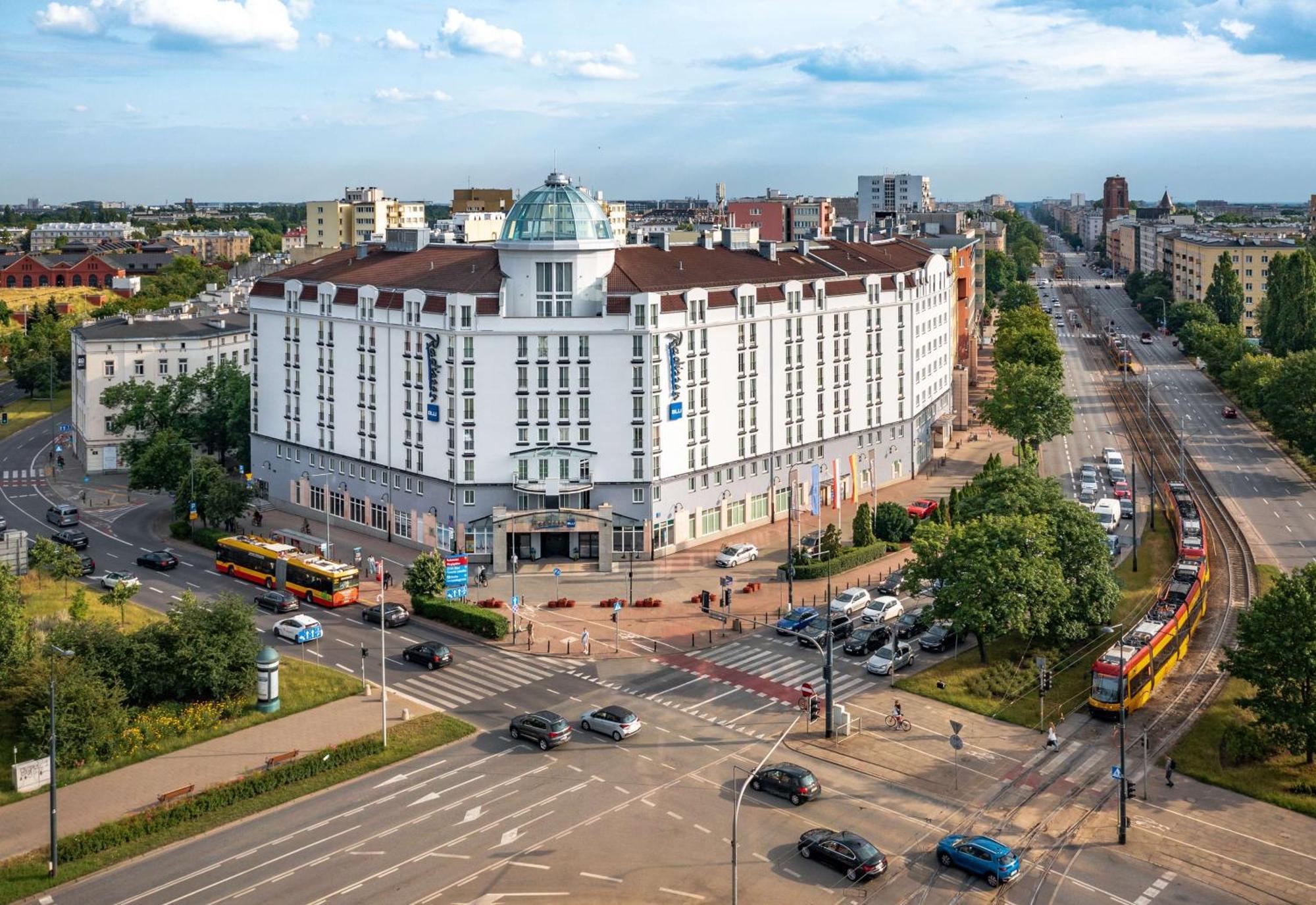 Radisson Blu Sobieski Hotel Warschau Buitenkant foto The photo shows a large, modern building with a distinctive architectural design, featuring a rounded dome on top. It's situated at a crossroads, surrounded by greenery and several roads. In front of the building, there are a few trams visible, indic